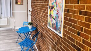 Une pièce avec un mur en briques, un tableau abstrait aux couleurs vives, une table pliante bleue avec des chaises, et une fenêtre ornée de rideaux blancs.
