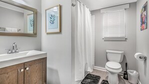 Une salle de bain avec un lavabo blanc, des toilettes, un rideau de douche et une fenêtre avec des stores.