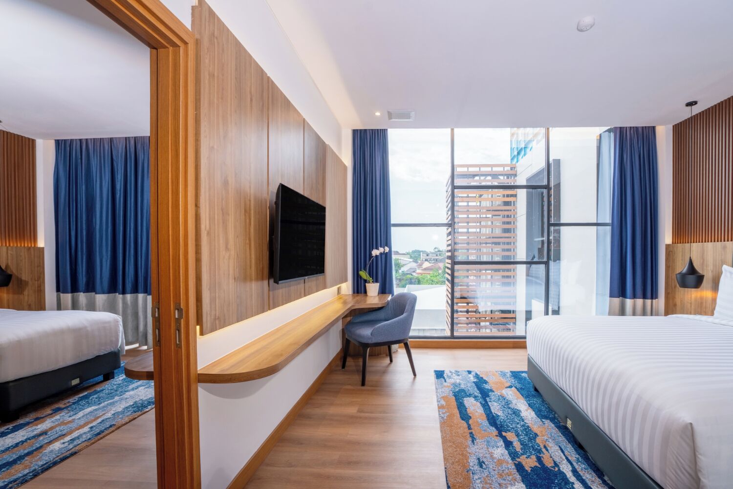 A modern hotel room with a large bed, a desk with a chair, a TV mounted on the wall, and a balcony with a view of the city.