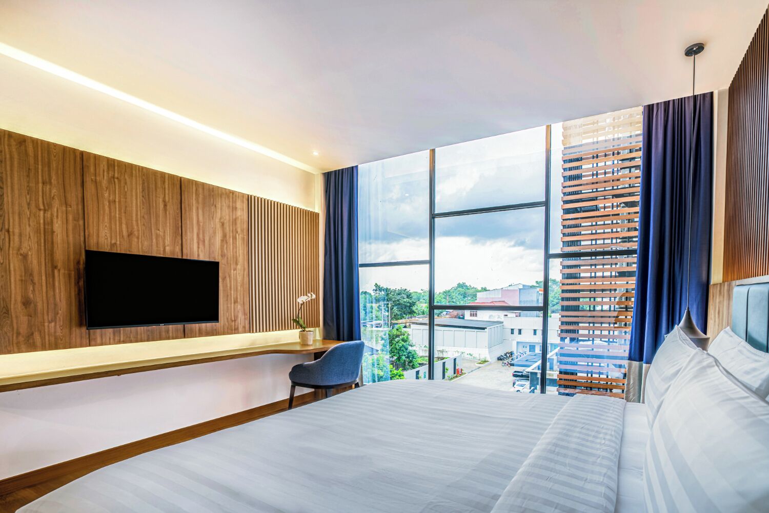 Habitación de hotel moderna con una cama grande, un televisor de pantalla plana montado en una pared de paneles de madera, un área de descanso con una silla y un ventanal con vistas a la ciudad.