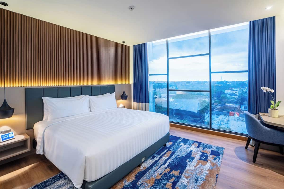 Una habitación de hotel moderna con una cama grande, un escritorio y vistas a la ciudad a través de ventanales de gran altura.