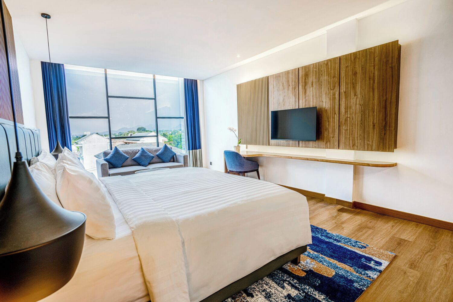 Una habitación de hotel moderna con una cama grande, un televisor montado en una pared de paneles de madera y vistas al exterior a través de amplios ventanales.
