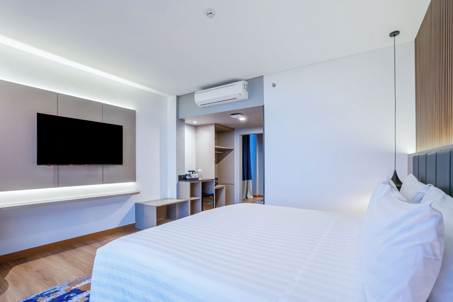 A modern hotel room with a large bed, a flat-screen TV mounted on the wall, a built-in wardrobe, and an air conditioning unit.