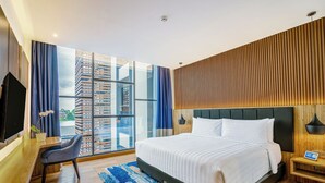Ein modernes Hotelzimmer mit einem großen Bett, einem Schreibtisch und Blick auf eine Skyline durch raumhohe Fenster.