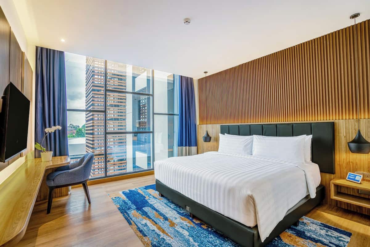 Habitación de hotel moderna con una cama grande, un escritorio y vistas a un paisaje urbano a través de ventanas de gran altura.