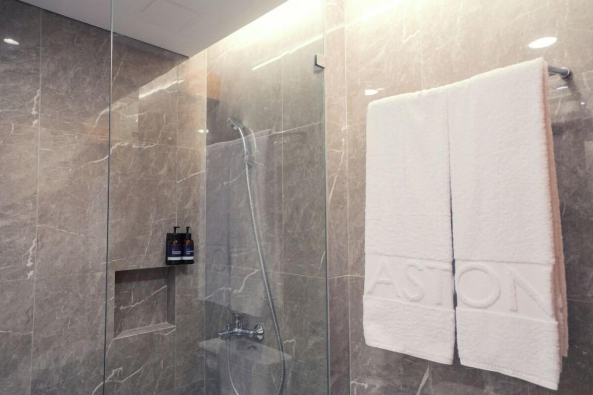 A modern bathroom with a glass shower enclosure, a marble wall, and a towel rack with a towel labeled "ASTON".