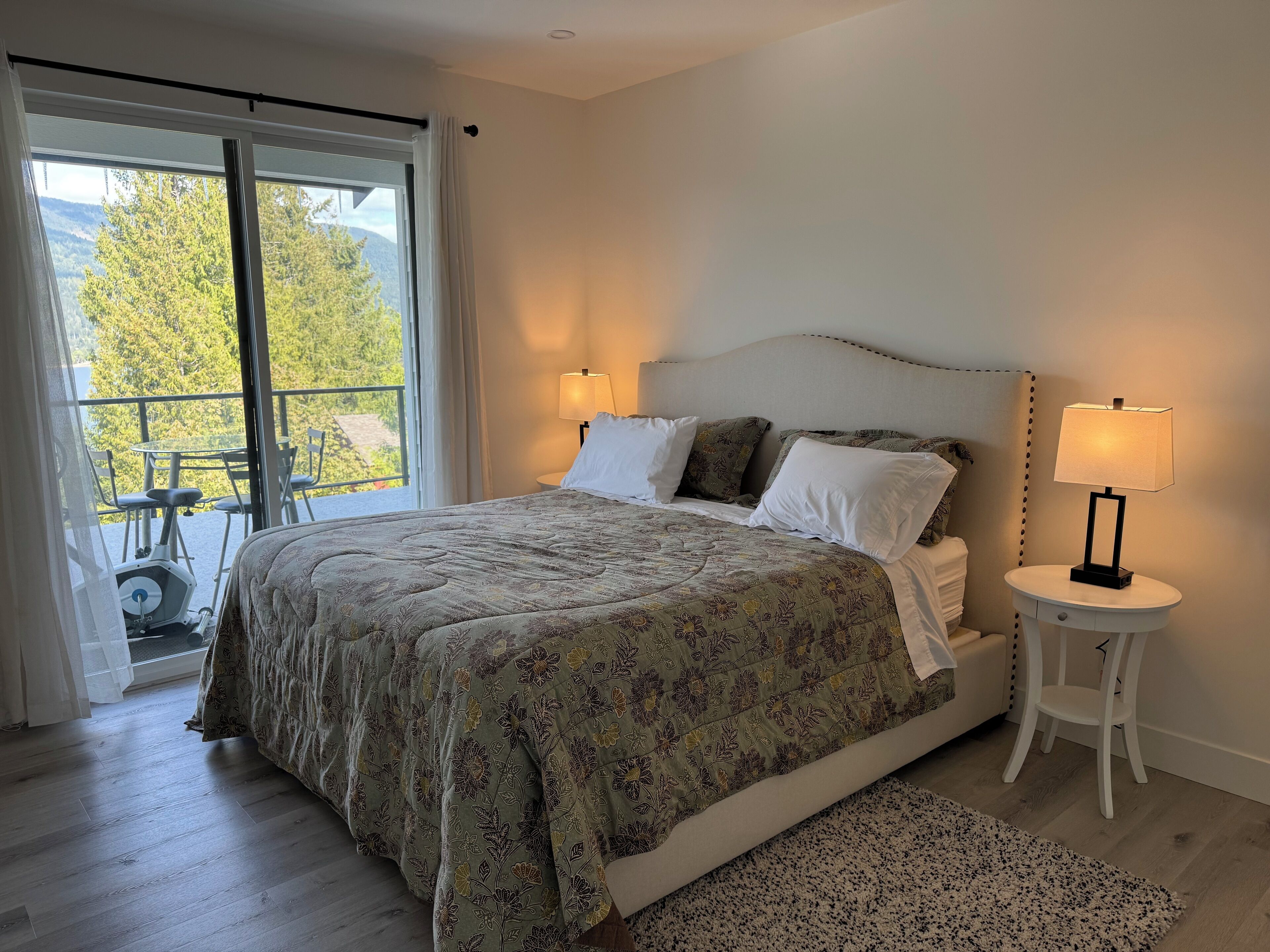 A bedroom with a bed, two bedside lamps, a small round table, and a view of a balcony with outdoor furniture.