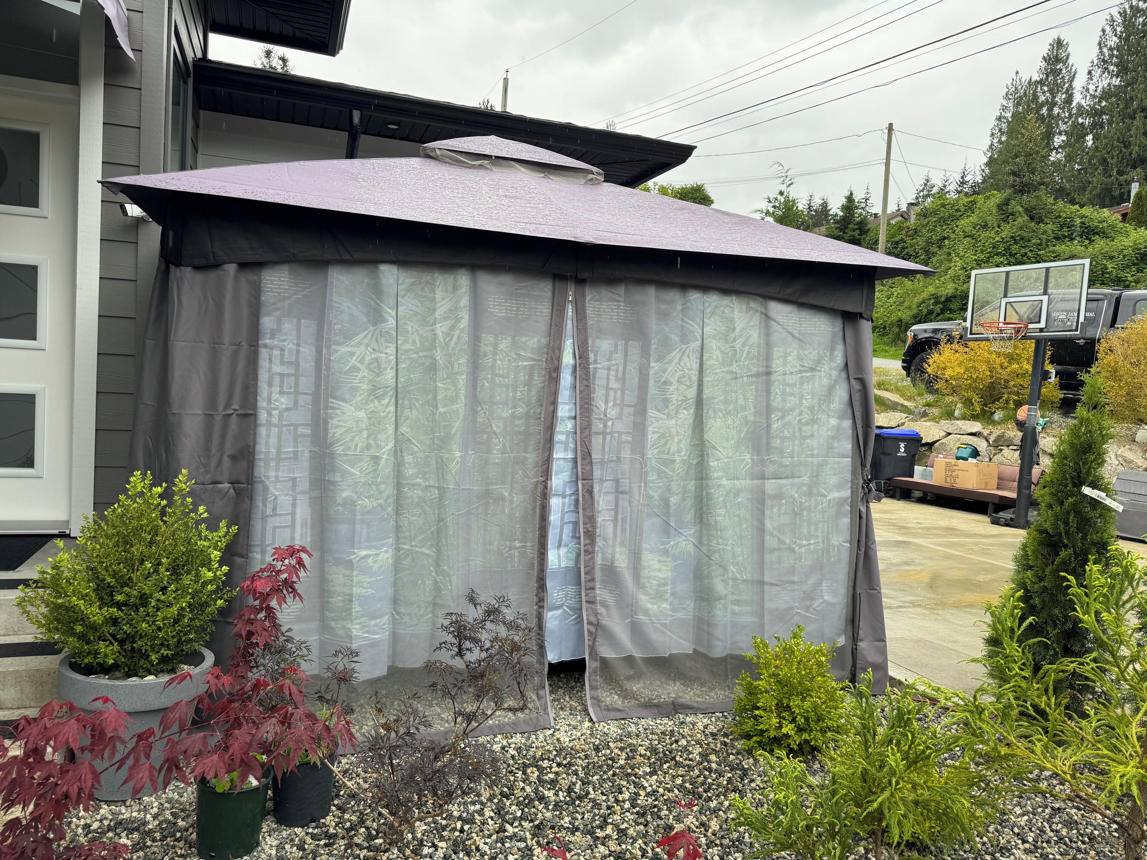 A covered outdoor area with a basketball hoop, surrounded by potted plants and a vehicle in the background.