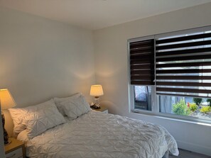 Une chambre à coucher avec un lit, deux lampes de chevet, une fenêtre avec des stores et une vue sur la verdure à l’extérieur.