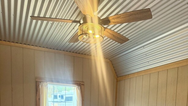 Une chambre avec un lit simple, des murs en bois, un plafond en pente avec un ventilateur de plafond et une fenêtre avec des rideaux.