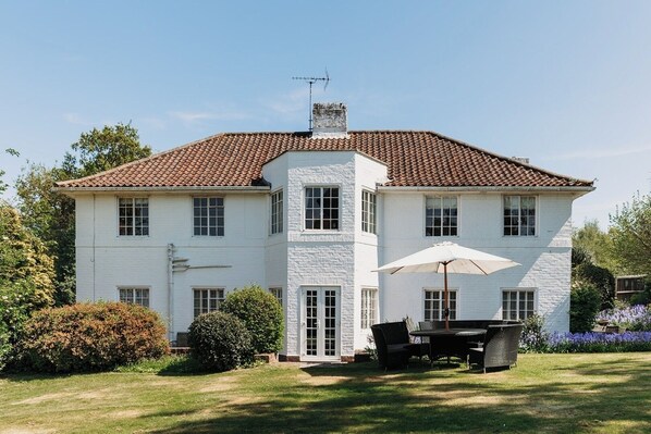 Ein zweistöckiges Haus mit Ziegeldach, weißen Außenwänden und einem Schornstein. Vor dem Haus befindet sich eine Terrasse mit Tisch und Stühlen, und ein Garten mit Büschen und Blumen.