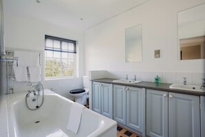 Ein modernes Badezimmer mit einer großen Badewanne, einer Toilette und einem Waschbecken. Ein Fenster mit Jalousien, ein Spiegel über dem Waschbecken und ein Handtuchhalter sind vorhanden.