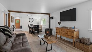 A spacious living room with a large sofa, a wooden dining table with chairs, a built-in wood-burning stove, and a large wall-mounted TV.