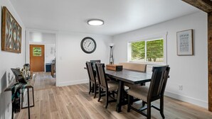A dining area with a wooden table and chairs, a wall clock, and a window with blinds.