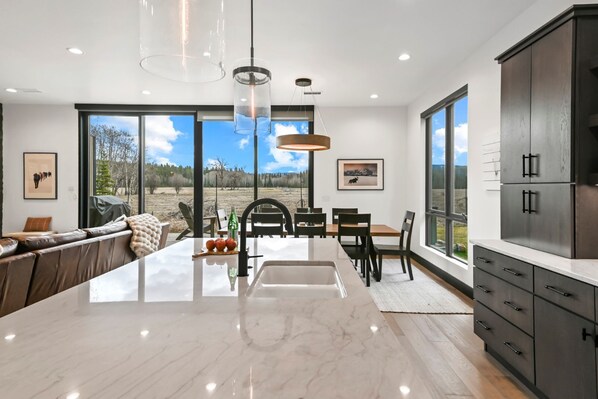 A modern kitchen with a large island, dark cabinetry, and large windows offering a view of the outdoors.