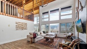 A spacious living room with a wooden ceiling, large windows, a sectional sofa, a coffee table, and a fireplace. - White Mtn Lux Retreat - Hot Tub, Sauna & Game Room (Bartlett)