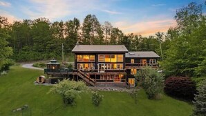 A two-story house with a deck, surrounded by a well-maintained lawn and trees. - White Mtn Lux Retreat - Hot Tub, Sauna & Game Room (Bartlett)