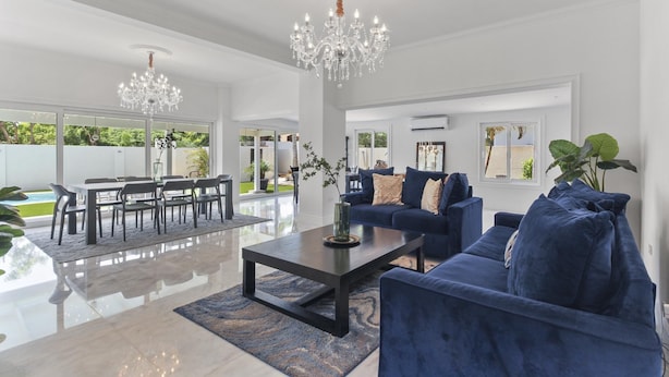 Ein geräumiges Wohnzimmer mit einer großen blauen Couch, einem Couchtisch und einem Kronleuchter. Es gibt einen Essbereich mit Tisch und Stühlen und einen Blick ins Freie durch große Fenster.