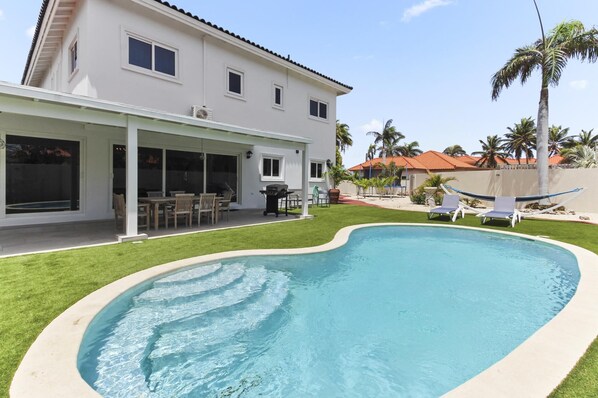 Ein Haus mit Swimmingpool, überdachter Terrasse und Rasen mit Palmen.