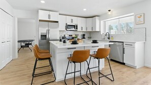 A modern kitchen with white cabinetry, a central island with stools, stainless steel appliances, and a window with blinds.