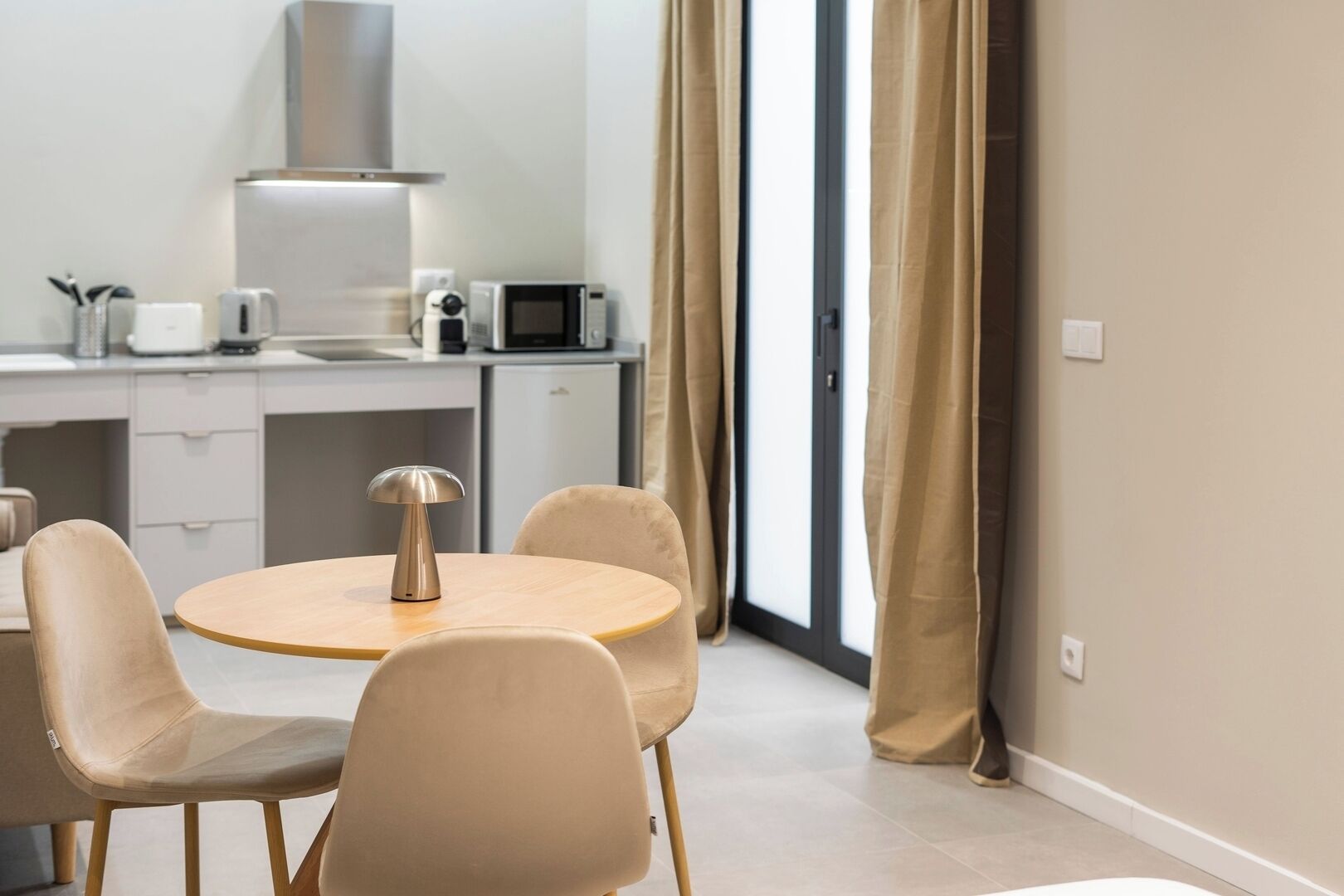 Ein stilvolles, helles Zimmer mit einem runden Esstisch, beigen Stühlen, einer kleinen Küchenzeile mit einer Mikrowelle und einem großen Fenster mit Vorhängen.
