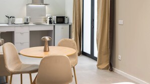 Ein stilvolles, helles Zimmer mit einem runden Esstisch, beigen Stühlen, einer kleinen Küchenzeile mit einer Mikrowelle und einem großen Fenster mit Vorhängen.