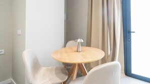 Une petite table ronde en bois, dotée d’un pied en métal au design moderne, est entourée de deux chaises blanches. Une fenêtre avec des rideaux de couleur beige est visible à l’arrière-plan.