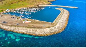An aerial view of a marina with boats docked, surrounded by a rocky breakwater and a grassy landscape.