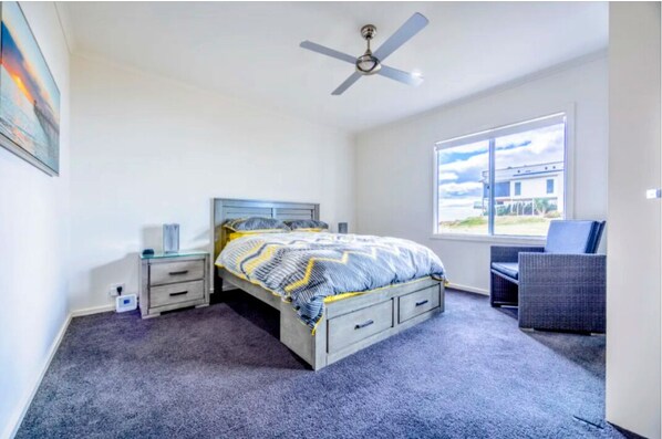 Un dormitorio con cama, mesita de noche, silla, ventana y ventilador de techo.