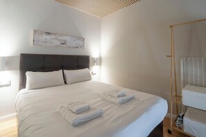 Un lit bien fait, avec du linge de lit et des serviettes blancs, une tête de lit sombre et un simple luminaire fixé au mur.