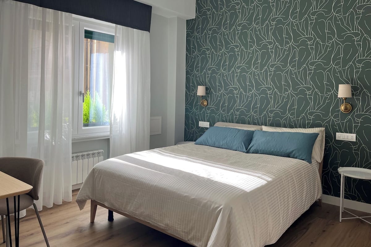 A bedroom with a large bed, a window with sheer curtains, a bedside table with a lamp, and a wall with a leafy pattern.