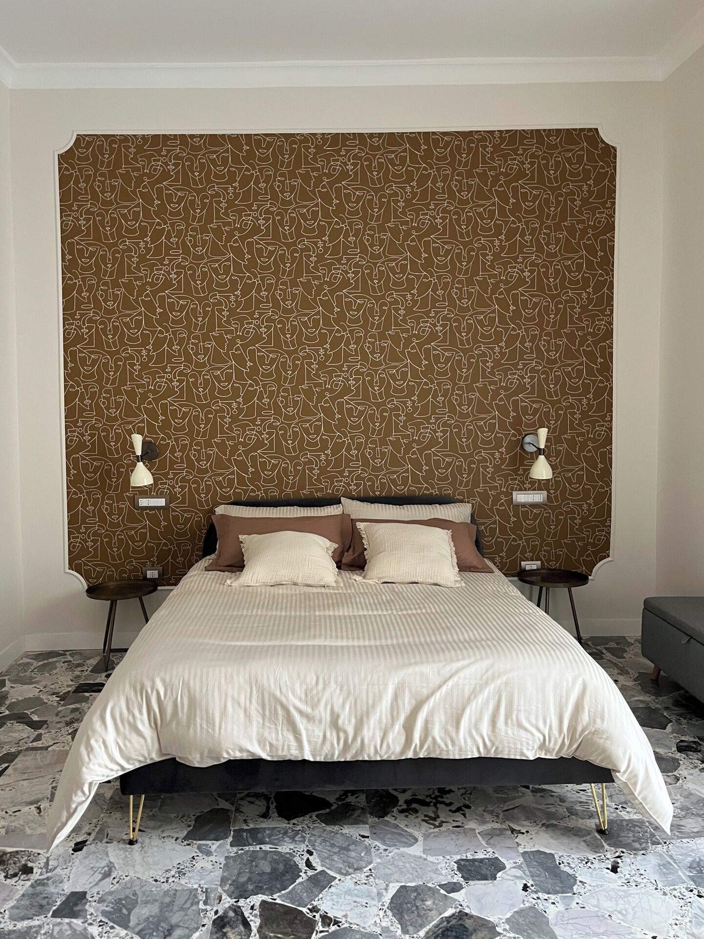 Une chambre à coucher avec une tête de lit à motifs, un lit recouvert d’une couvre-lit à rayures, deux tables de chevet avec des lampes et un sol carrelé en pierre.