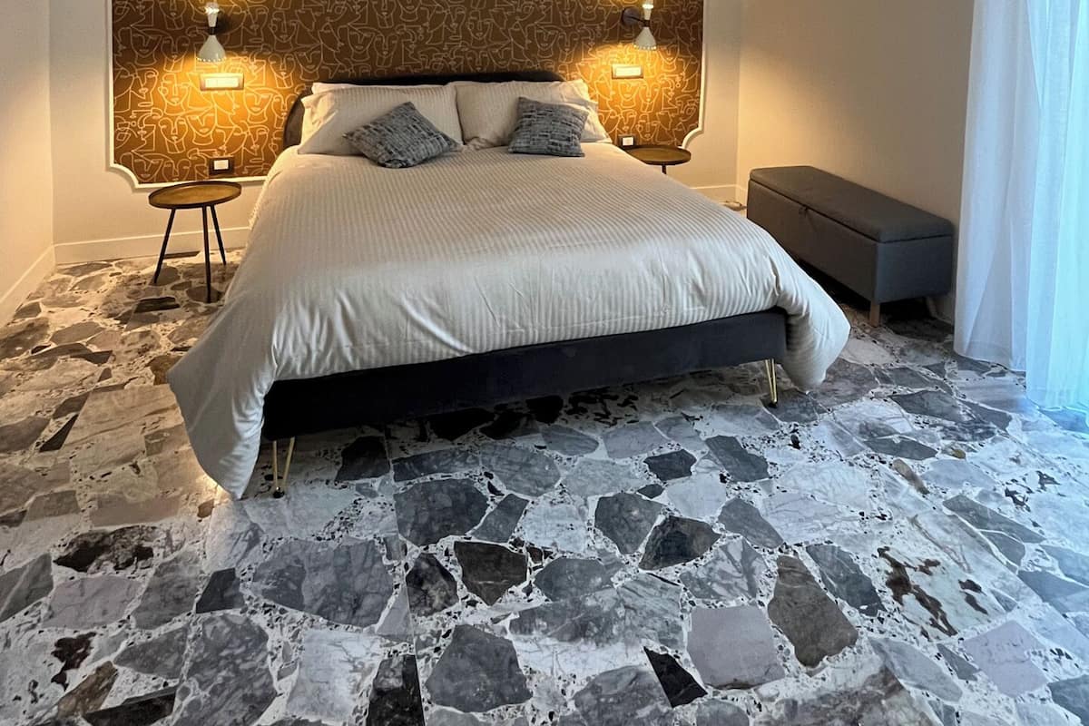 A bedroom with a patterned headboard, two bedside lamps, a bed with pillows, a bench, and a stone floor.