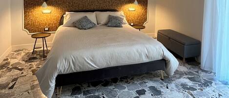 A bedroom with a patterned headboard, two bedside lamps, a bed with pillows, a bench, and a stone floor.