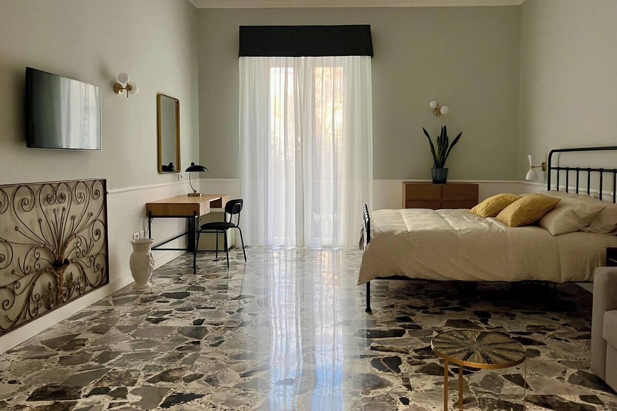 A bedroom with a bed, a desk, a chair, a TV, and a large window with curtains.