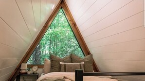 Ein Schlafzimmer mit schrägem Dach, einem Bett mit Kissen, einem Nachttisch mit Lampe und Blick auf einen Wald durch ein großes, dreieckiges Fenster.