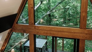 Eine dreieckige Holzhütte mit großen Fenstern, die einen Blick auf einen Wald bieten. Im Inneren befindet sich ein Tisch und Stühle in Fenster Nähe.