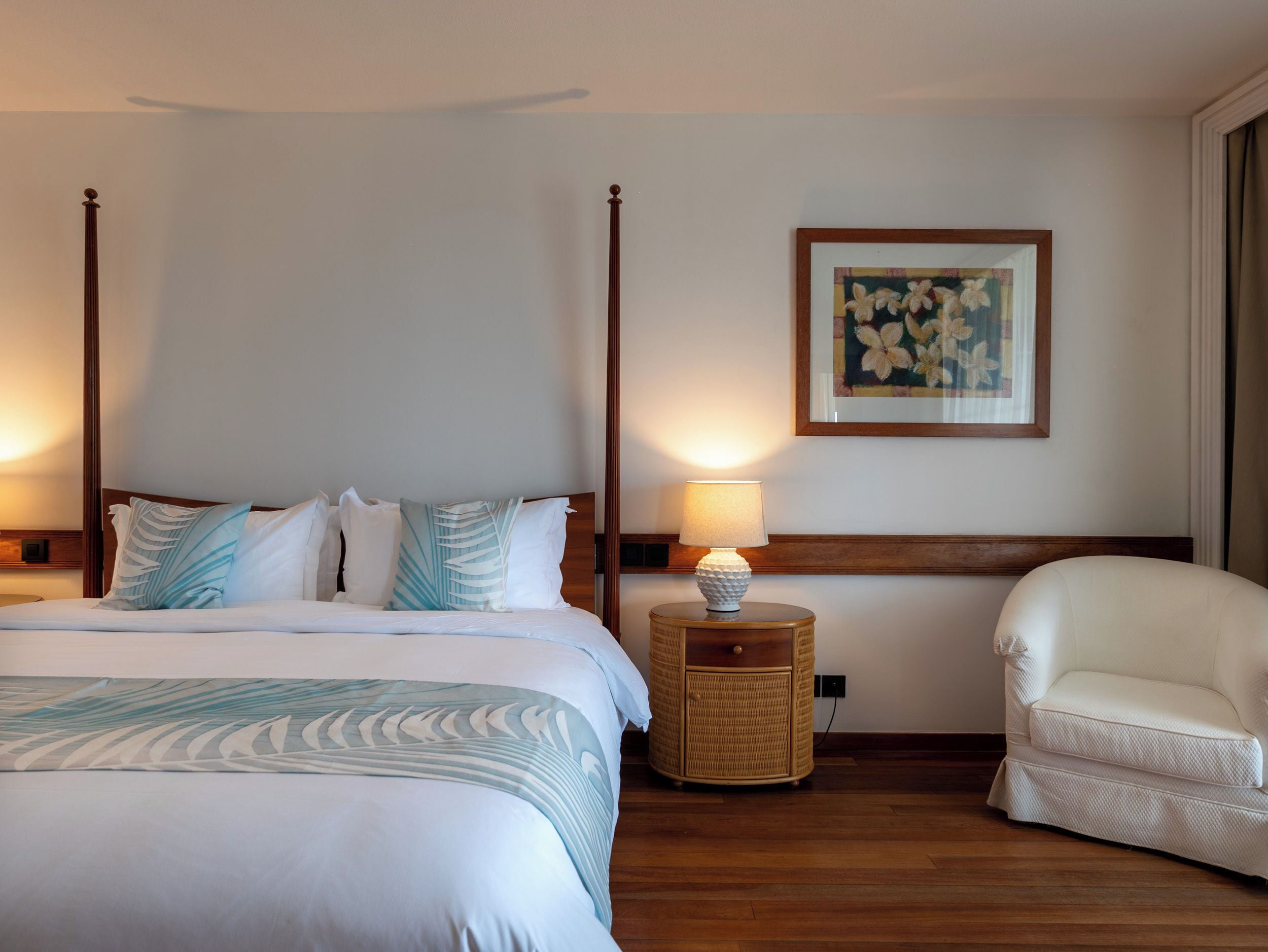 Una cama con dosel con un edredón blanco y azul estampado, una lámpara de noche, una mesita de noche de mimbre, un sillón blanco y un cuadro floral enmarcado en la pared.