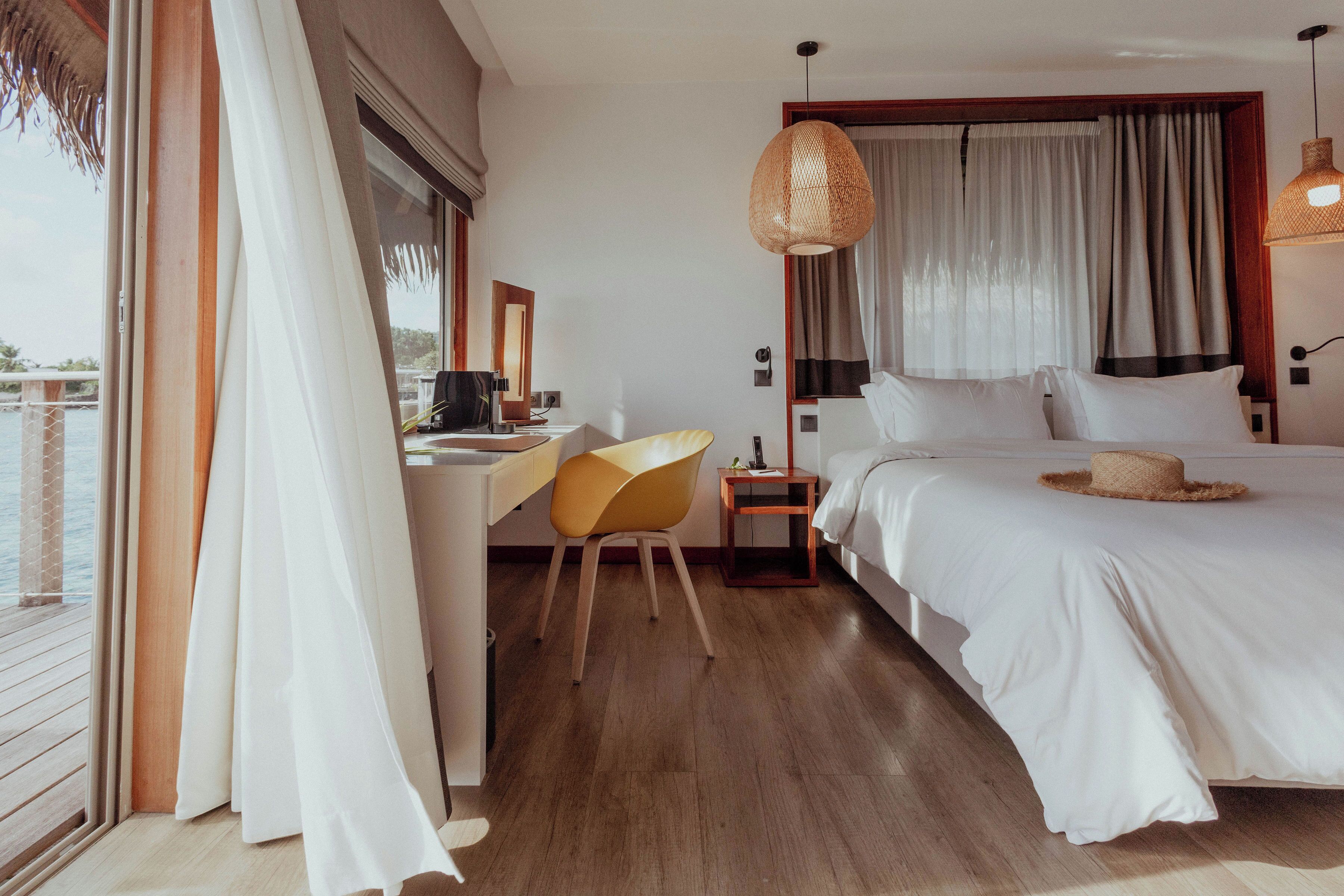 A bedroom with a bed, a desk, a chair, a nightstand, and a large window with curtains.