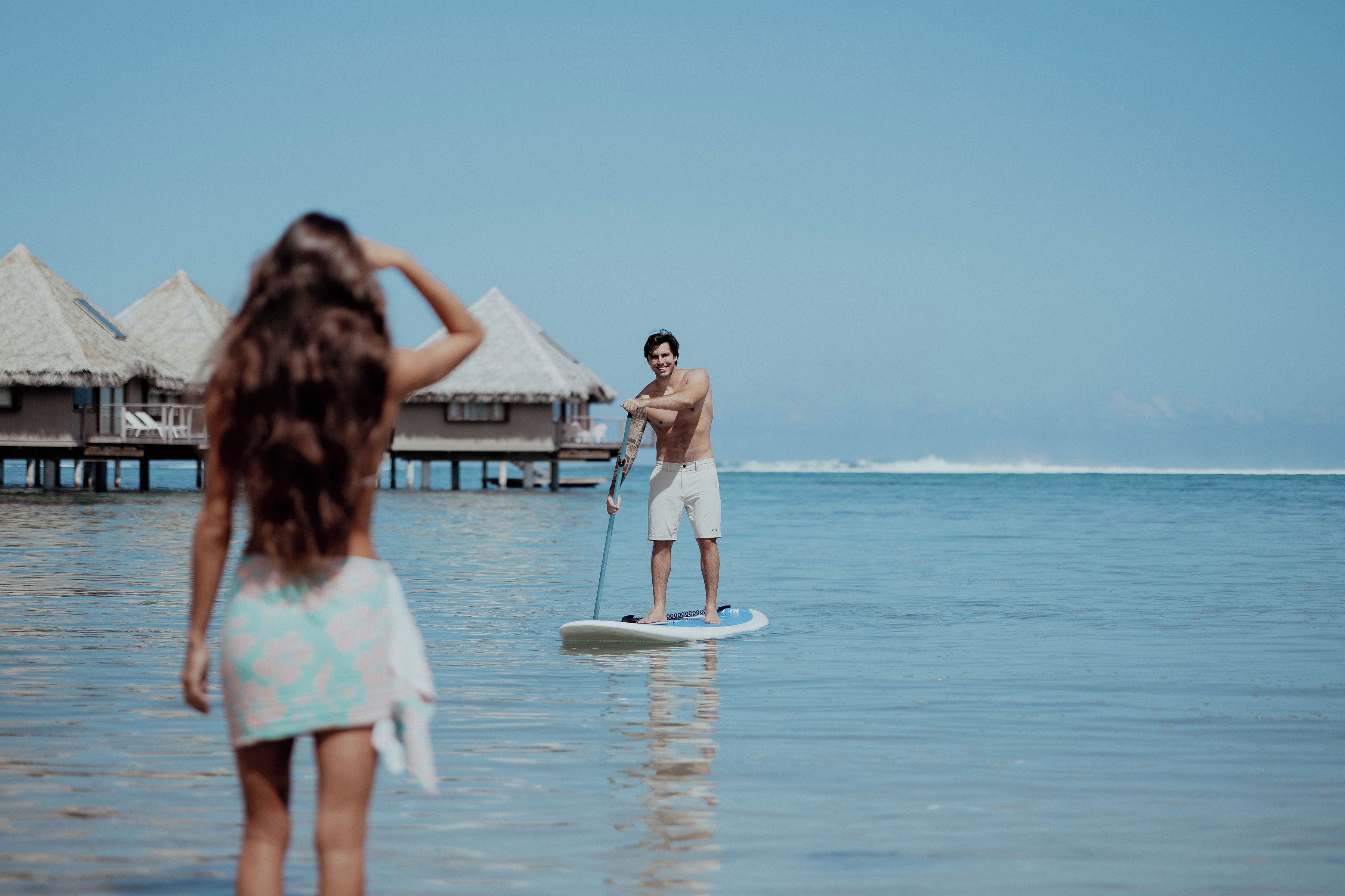 A person paddleboarding on calm ocean waters with overwater bungalows in the background.