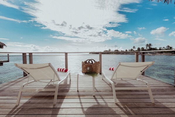 A wooden deck with two white lounge chairs and a wicker bag, overlooking a calm sea and palm trees.