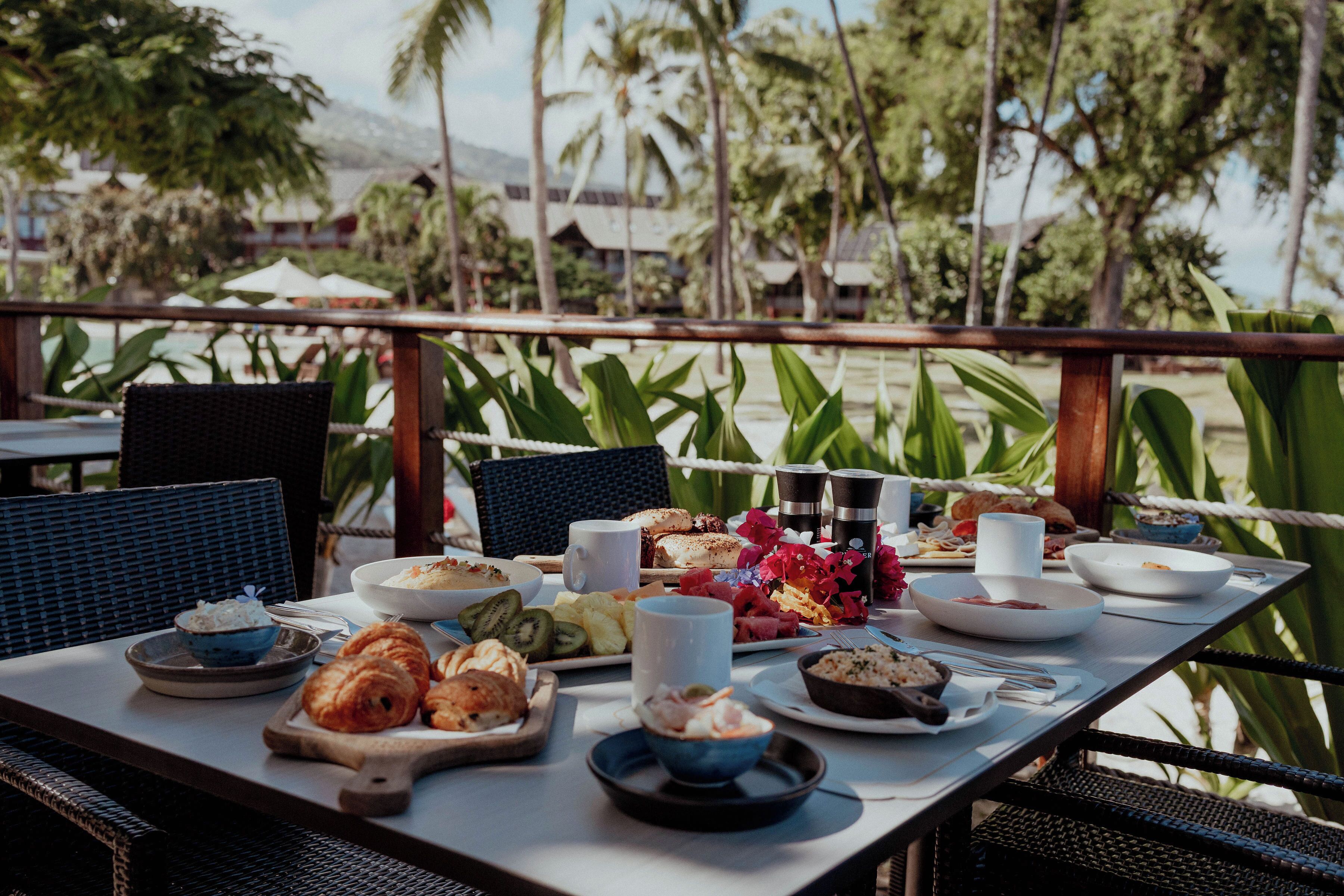 Une table dressée avec divers plats, notamment des croissants et des fruits, sur un balcon donnant sur une plage bordée de palmiers.