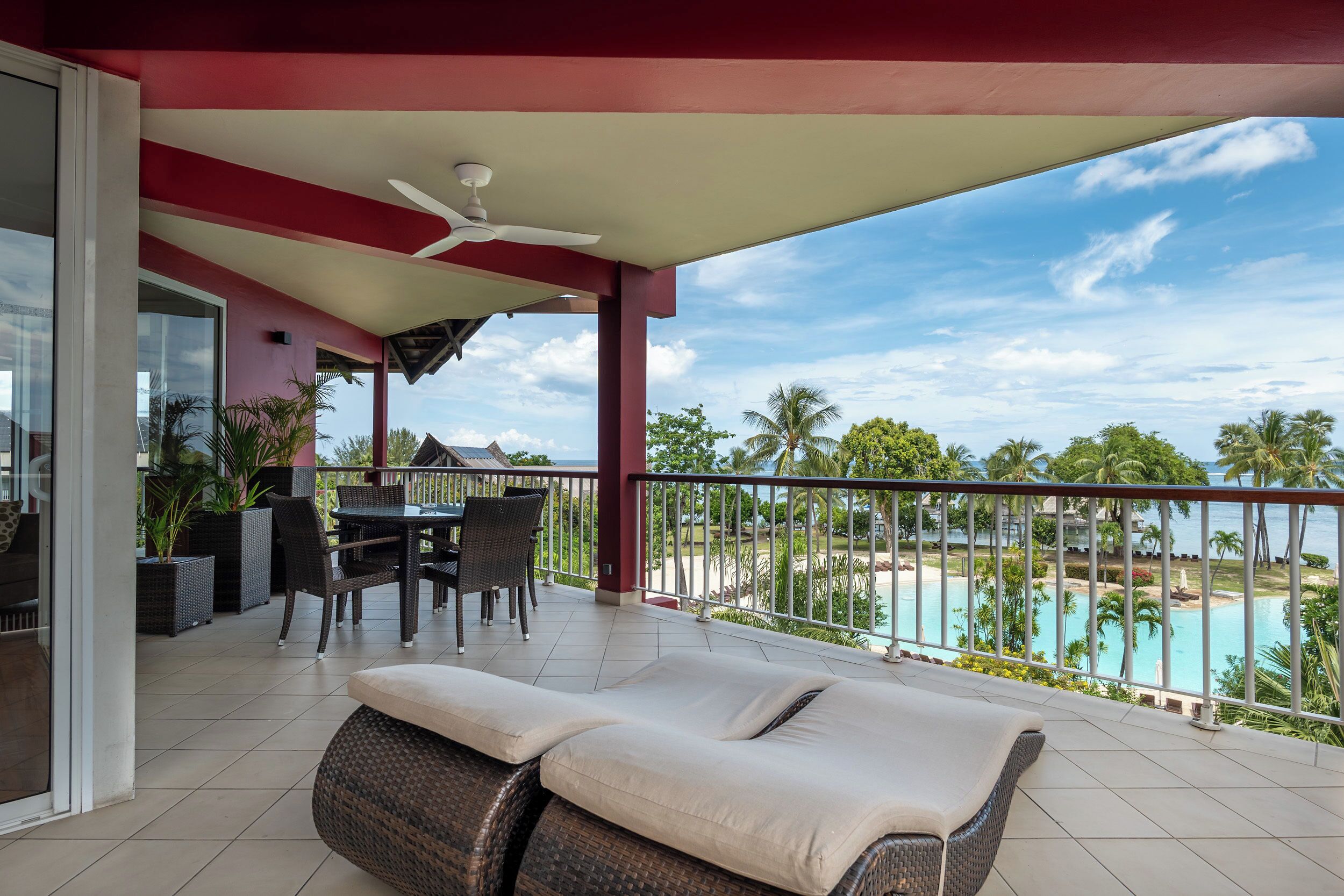 A balcony with a view of a pool and palm trees, a table with chairs, and a lounge area with cushions.