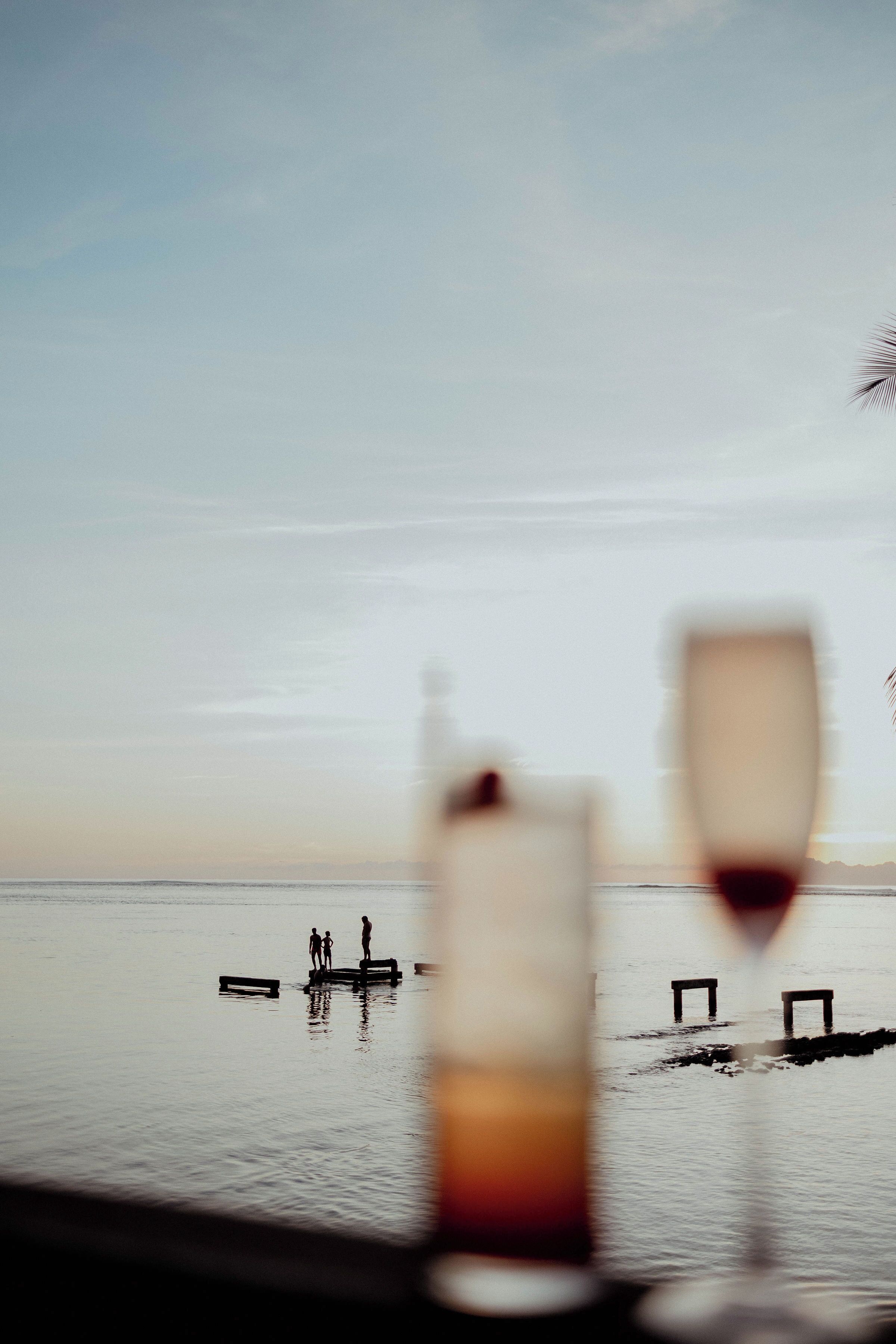 A view of a calm body of water with two people standing on a dock, and two glasses of wine in the foreground.