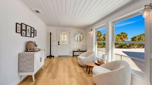 Una habitación amplia con suelo de madera, una puerta blanca con un vitral, un espejo redondo y vistas a la playa a través de grandes ventanales.