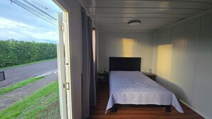 A small, single-bed room with a wooden floor, a bedside table, and a window overlooking a road and greenery.