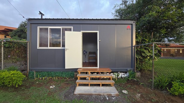 A small, single-story building with a grey exterior, a white door, and a small porch area with a bench.