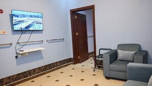 A waiting area with a television, a grey sofa, a small table, and a wheelchair. - Mawed Al Dyafa Serviced Apartments  (Riyadh)