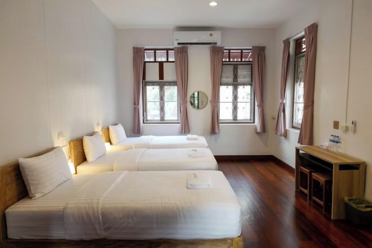 A hotel room with three beds, a wooden floor, a small table with a trash bin, and a wall-mounted air conditioner.