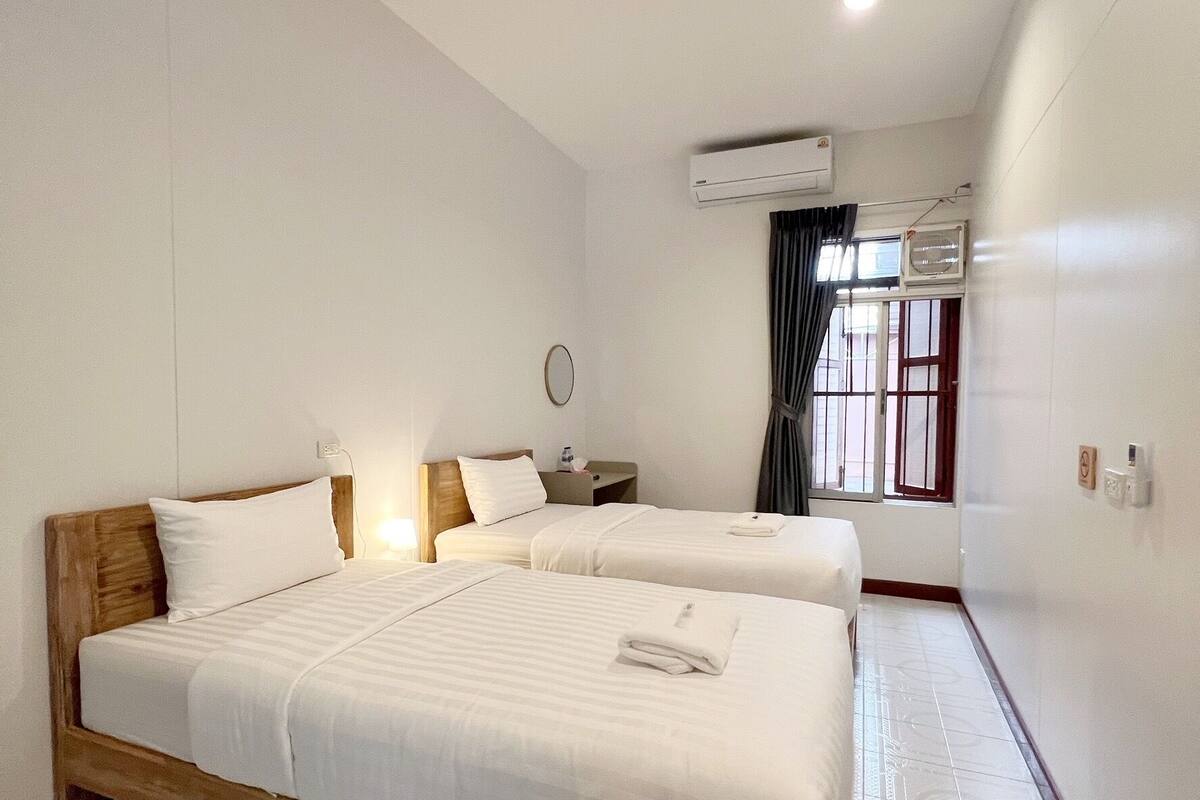 A hotel room with a large bed, a smaller bed, a bedside table, an air conditioning unit, a window with curtains, and a tiled floor.